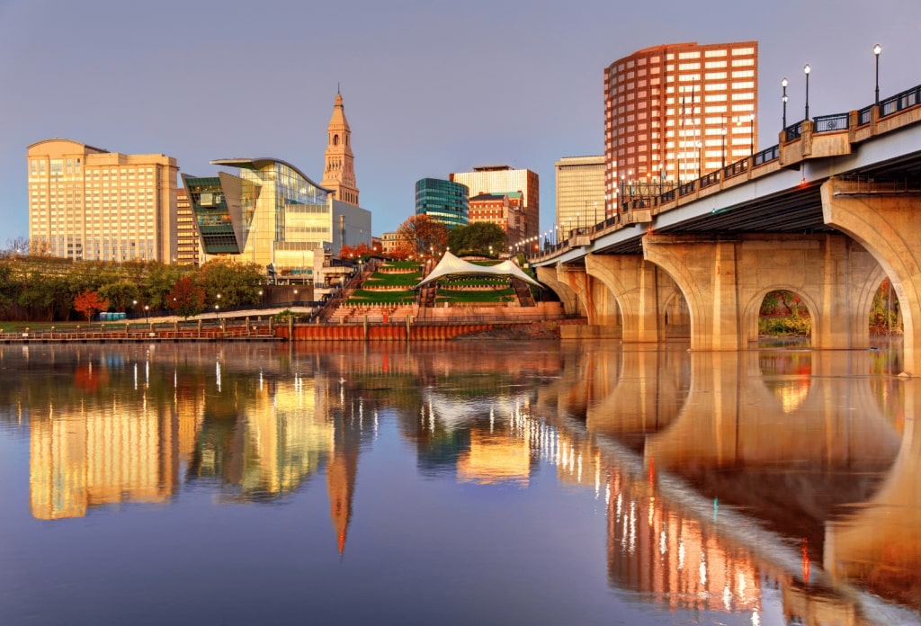 The Hartford, Connecticut skyline at sunset, featuring the riverfront, the historic Travelers Tower, and the Founders Bridge reflecting in the water.