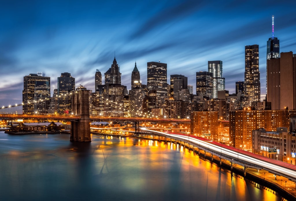 A scenic evening view of the Brooklyn Bridge with the illuminated New York City skyline and light trails from traffic in the background.
