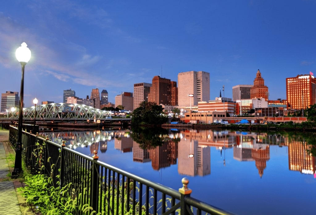 A tranquil evening view of a city skyline reflected in a calm river, featuring a pedestrian bridge and a well-lit waterfront walkway.