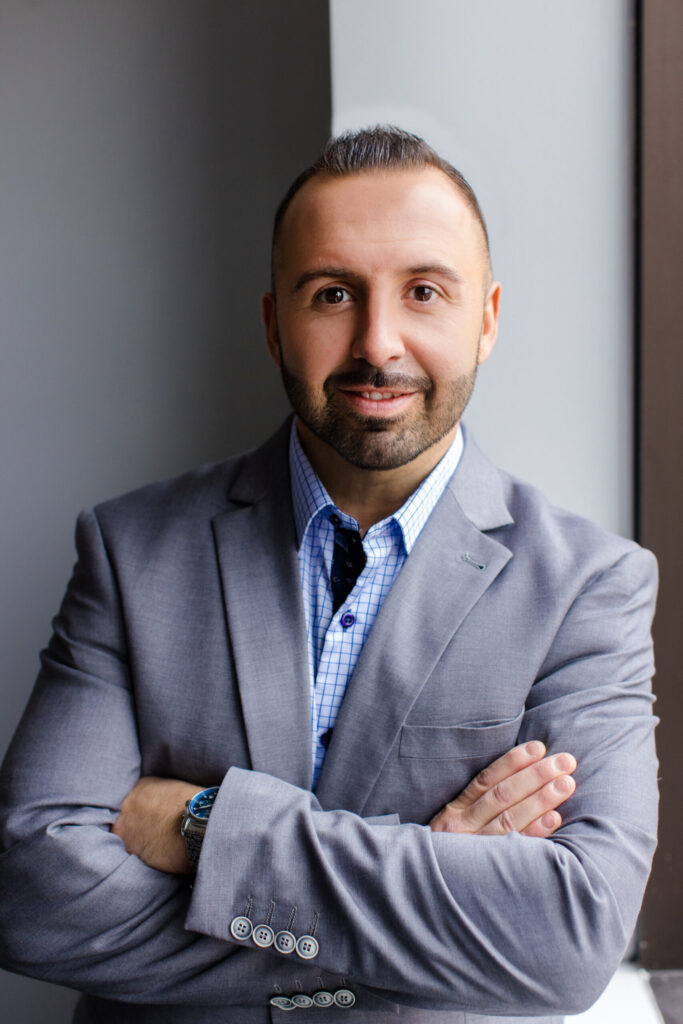 Shows a professional headshot of a smiling man with a beard, wearing a grey blazer over a blue and white checkered shirt, posing with his arms crossed.