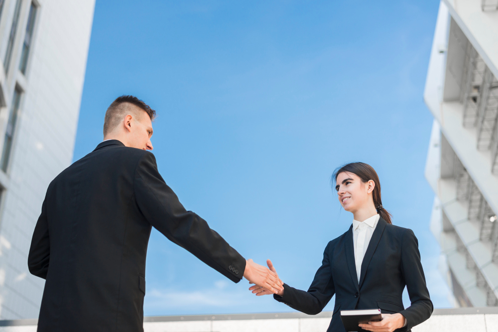 Depicts a man and a woman in formal business attire shaking hands outdoors, with modern architectural buildings in the background and a clear blue sky overhead.
