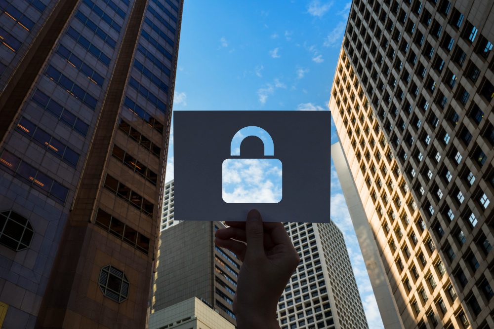 Depicts a person holding up a piece of paper with a cutout of a padlock symbol, positioned against a background of tall city skyscrapers and a blue sky.