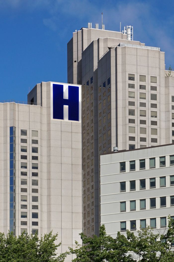 Features a large hospital complex, identified by a prominent blue "H" sign on its grey concrete facade.